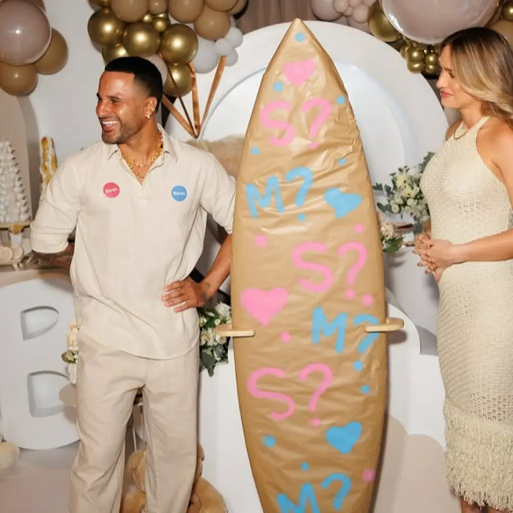 A man and a woman standing in an event space, with the man smiling and holding a decorated surfboard covered in pink and blue question marks and hearts, symbolizing a gender reveal celebration.
