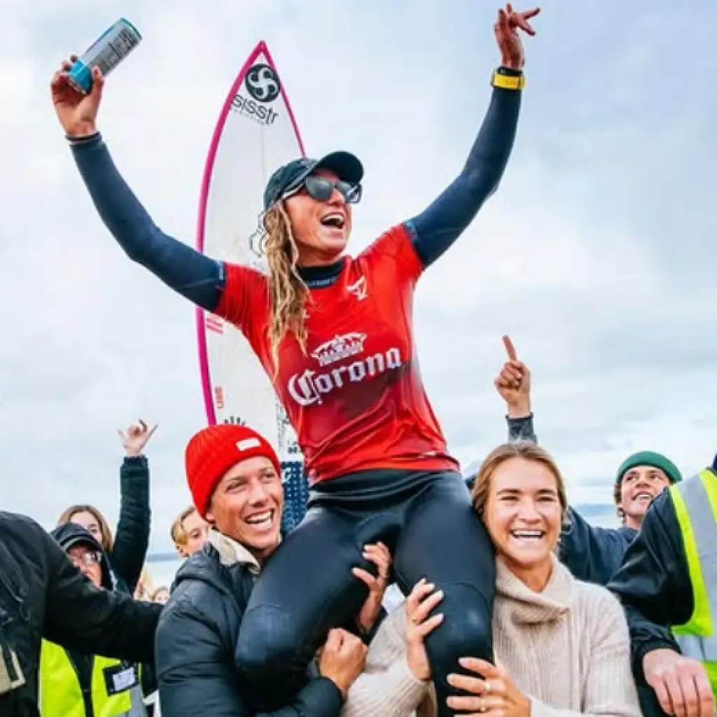 A joyful female surfer wearing a red jersey with the Corona logo is being lifted on the shoulders of two supporters while celebrating, with a surfboard and enthusiastic fans visible in the background.