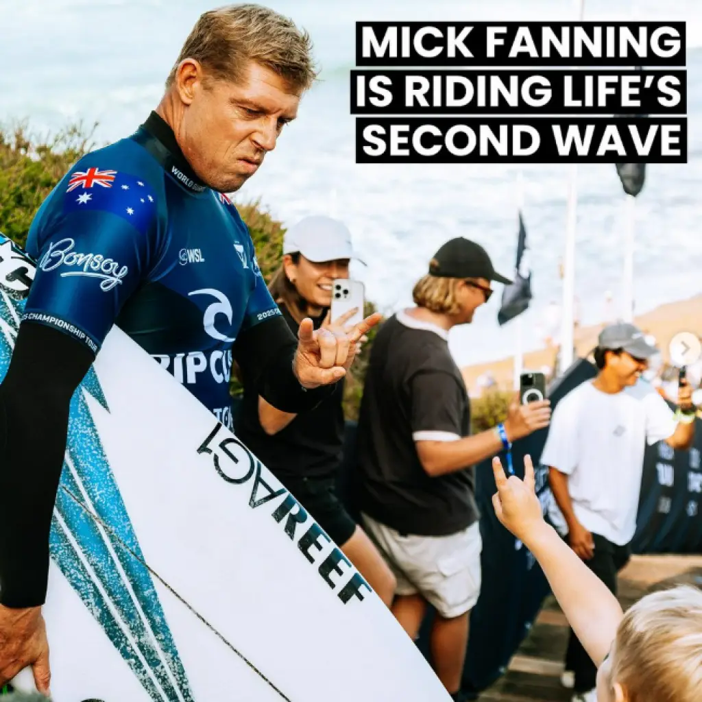 Mick Fanning, a professional surfer, poses with his surfboard while interacting with a fan, set against a crowd at Jeffreys Bay.